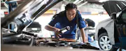 Car mechanic leans over opened car hood to work on vehicle.