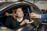 A woman sitting in a car smiles as someone hands her the keys.